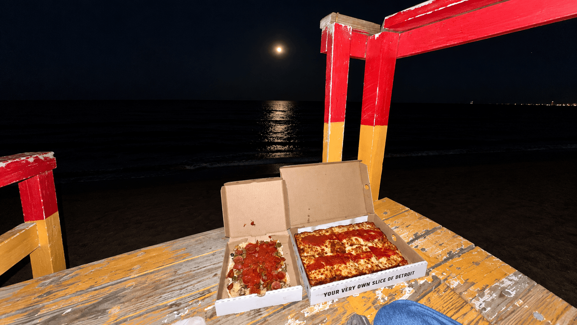 Two open pizza boxes beside the water at night under a full moon